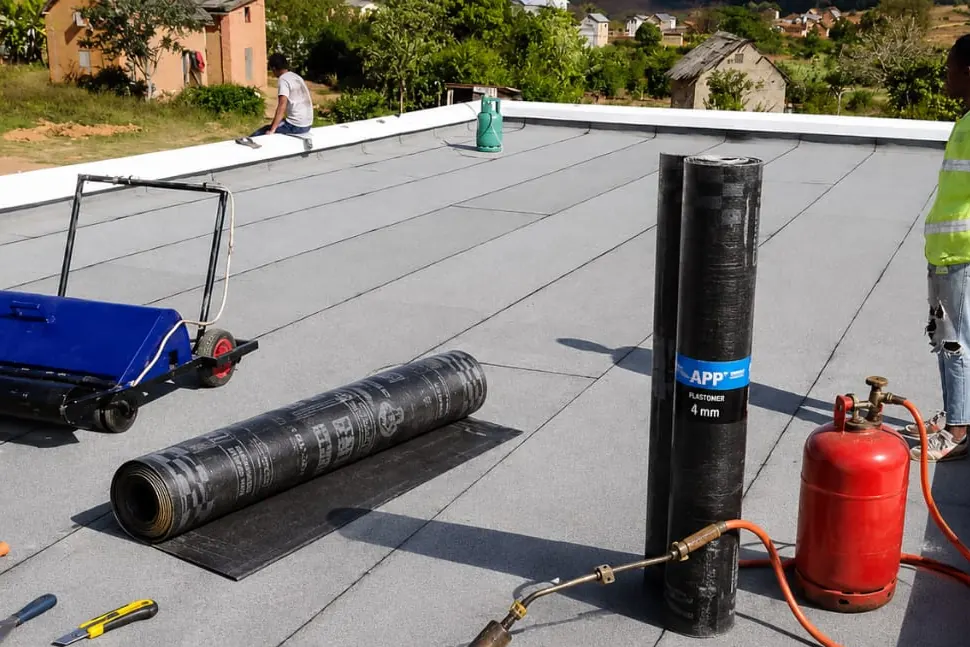 Rouleaux de membrane bitumineuse, chalumeau et bouteille de gaz sur une toiture-terrasse préparée.